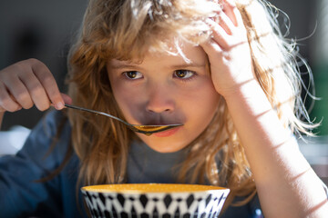 Unhappy sad child boy with spoon eats itself. Kid eating food on kitchen.