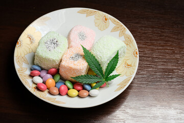 Cannabis dessert. Turkish delight with coconut flakes and colorful candy confetti, top view. Green marijuana leaves on white plate background. Layout of hemp food with CBD oil.