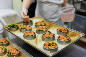 Female cook is filling group of tarts with salmon at restaurant kitchen background. Culinary process