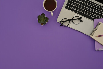 Top view of a purple desk with glasses on a laptop keyboard, a cup of coffee, a cactus and other office supplies in the copy space. Flat lay composition.