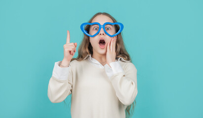 Surprised kid in funny heart-shaped glasses keeping finger raised blue background, eureka