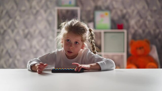 Smart little girl student with toy abacus does sums sitting at white table during mental mathematics lesson against Teddy bear closeup slow motion
