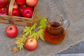 Glaskrug mit Apfelsaft vor einem Korb mit roten Äpfeln.
