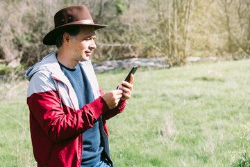 Entrepreneurial self-employed man wearing a hat, looking at his mobile phone while relaxing in the field, on a meadow. Concept of work, enjoy, relax, Internet, technology and connectivity.