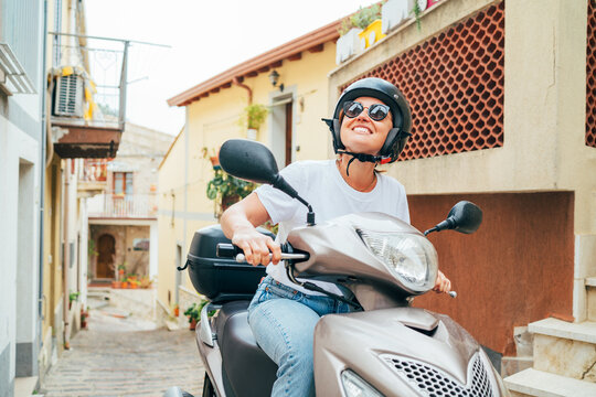 Cheerfully smiling woman in helmet and sunglasses fast riding the moto scooter on the narrow Sicilian old town streets. Happy Italian vacation and transportation concept.