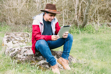 Obraz premium Entrepreneurial self-employed man wearing hat, looking at his tablet while relaxing in the field, sitting on a stone. Concept of work, enjoy, relax, Internet, technology and connectivity.