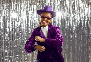 Funny extravagant young african american woman having fun dancing on shiny silver background. Woman in purple velvet jacket, hat in sunglasses having fun on background of fringed foil curtains.