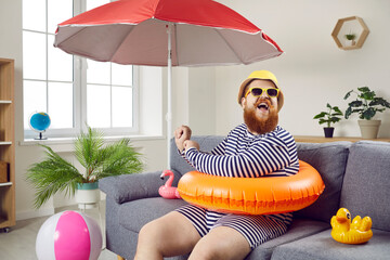 Summer party at home. Funny chubby man having fun sitting on sofa in living room with inflatable swimming circle. Humorous man has absurd vacation under beach umbrella on improvised home beach. - obrazy, fototapety, plakaty