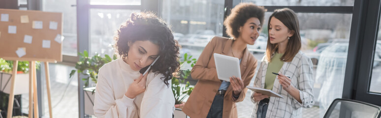 Arabian businesswoman talking on cellphone near multiethnic colleagues with digital tablet in office, banner.