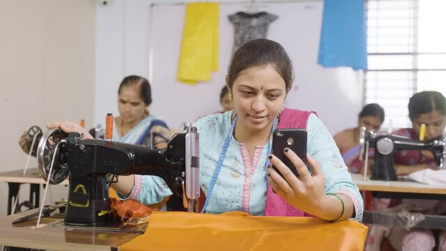 Women tailor working by watching mobile phone in front of sewing machine at garments - concept of technology, learning and employment