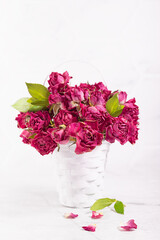 Elegant pink dry roses bouquet with fresh green leaves in white basket in soft light white kitchen interior as holiday decor, copy space,  vertical.