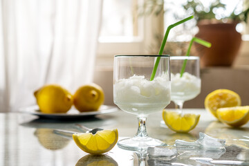 Lemon sorbet in two glasses with lemons, ice cubes and drinking straws on grey background and window backside
