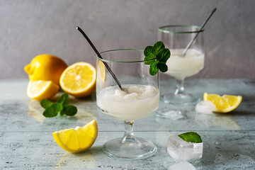 Lemon sorbet in two glasses with lemons, ice cubes, tea spoons, mint leaves on grey background