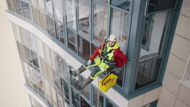 Industrial climber fools around hanging on safety ropes against windows of skyscraper. Worker relaxes during work process on height view from above
