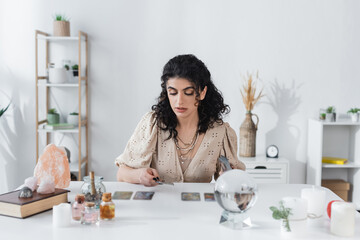 Gypsy fortune teller holding tarot cards near candles and witchcraft supplies on table.