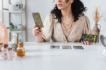 KYIV, UKRAINE - FEBRUARY 23, 2022: Cropped view of fortune teller holding tarot cards near crystal at home. - obrazy, fototapety, plakaty