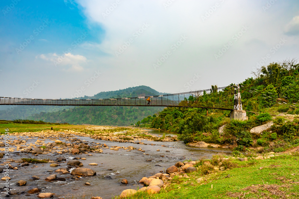 Wall mural isolated iron suspension bridge over flowing river with mountain and blue sky background at morning