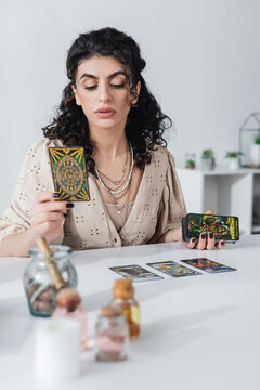 KYIV, UKRAINE - FEBRUARY 23, 2022: Gypsy fortune teller holding tarot cards near blurred witchcraft supplies at home.