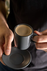A man's hand holds a cup of hot espresso coffee against the background of a black T-shirt.