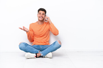 Young man sitting on the floor isolated on white background keeping a conversation with the mobile phone with someone