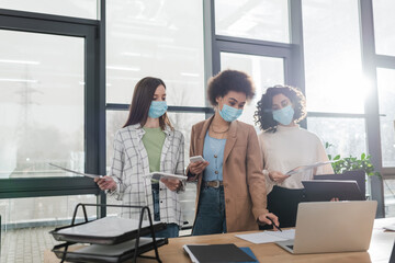 Multiethnic businesswomen in medical masks holding devices and working with papers in office.