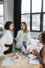 Cheerful businesswoman talking to multiethnic colleagues near devices in office.