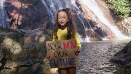 Child girl holding Your now, our future, in the background of a waterfall. Poster showing a sign protesting against plastic pollution in the forest. The concept of World Environment Day. Zero waste. 