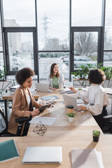 Positive interracial businesswomen talking near devices and papers on table in office.