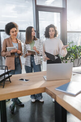 Arabian businesswoman holding documents and talking to interracial colleagues near blurred devices in office.