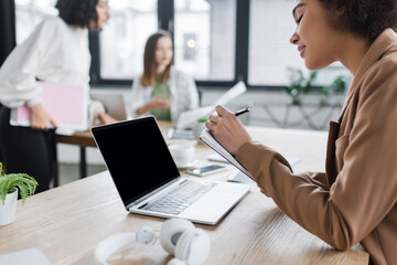 Young african american businesswoman writing on notebook near laptop with blank screen in office.