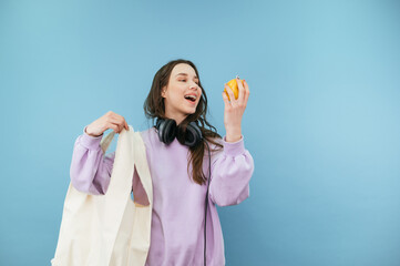Attractive woman with an eco bag in her hands stands on a blue background with an apple in her hand and looks at the fruit with a smile on her face.