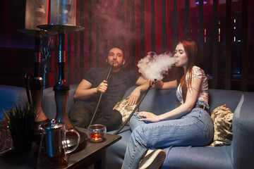 a young woman and a man are talking in a hookah room, both are smoking a hookah, releasing a large...