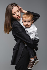 young woman fixing hair while holding son grimacing at camera isolated on grey.