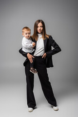 businesswoman in black suit standing with toddler son on grey.