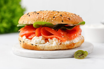 Bagel sandwich with salmon, cream cheese and green leaves on marble board and white background. Burger with salmon