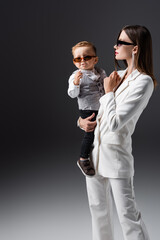 elegant woman holding toddler son wearing sunglasses on grey.