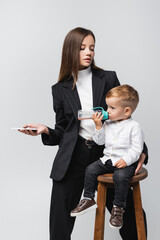 woman in black suit holding smartphone near son drinking from baby bottle isolated on grey.