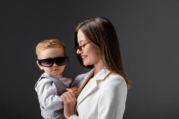 toddler boy in trendy sunglasses looking at camera near happy mom isolated on grey.