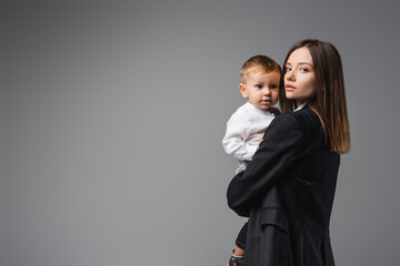 woman in black blazer holding toddler son and looking at camera on grey.