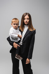 businesswoman in suit holding toddler kid and looking at camera isolated on grey.