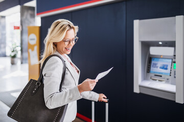 A beautiful blonde middle-aged businesswoman stands at a train station with a ticket in her hand.
