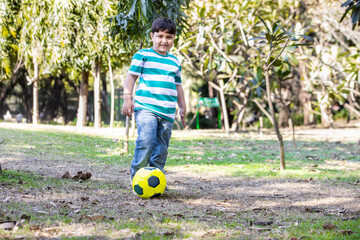 Young indian boy kid play soccer in the park garden outdoor looking at camera. smiling asian sporty male child kick yellow football, copy space.