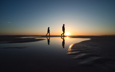 Silhouette von zwei Personen die während des Sonnenuntergangs am Strand spazieren gehen - obrazy, fototapety, plakaty