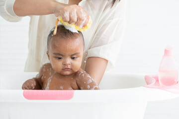 Cute African newborn baby bathing in bathtub with soap bubbles on head and body. Mother use sponge...