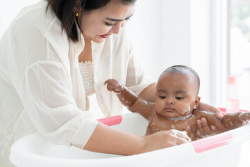 Adorable African newborn baby bathing in bathtub. Asian young mother use sponge with shower gel...