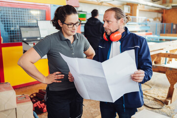 two carpenters stand in front of a cnc machine and look at a plan