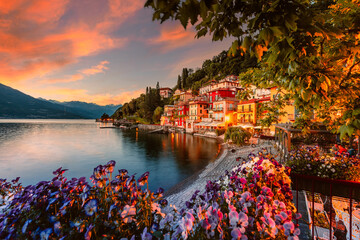 Village of Varenna on Lake Como at sunset with illuminated houses and colorful flowers in the foreground - obrazy, fototapety, plakaty