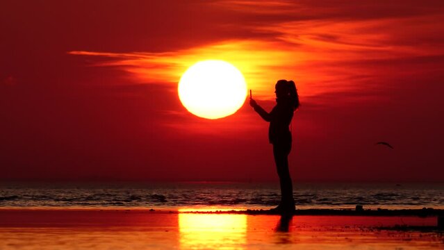 Silhouette of tourist woman, she take pictures using smartphone, big sun disc on background. Shallow place or sand bar at beach, cool autumn breeze from water, small waves on background