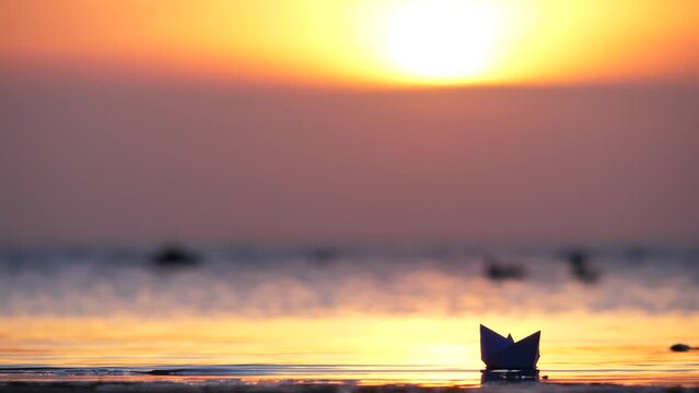 Male hand carefully holds paper boat and puts it on water, tiny toy boat sails away. Blurred sea and bright sunset seen on background, some birds stay on small waves