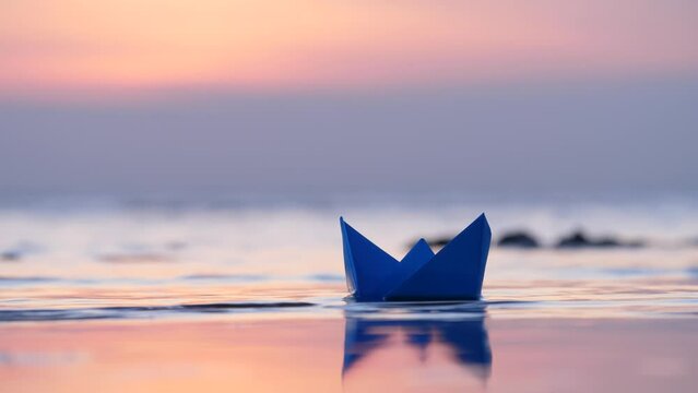 Little paper boat ran aground at evening hour, standing motionless in shallow water near shore. Small waves can be seen in blurred background, close up shot of toy ship in trouble